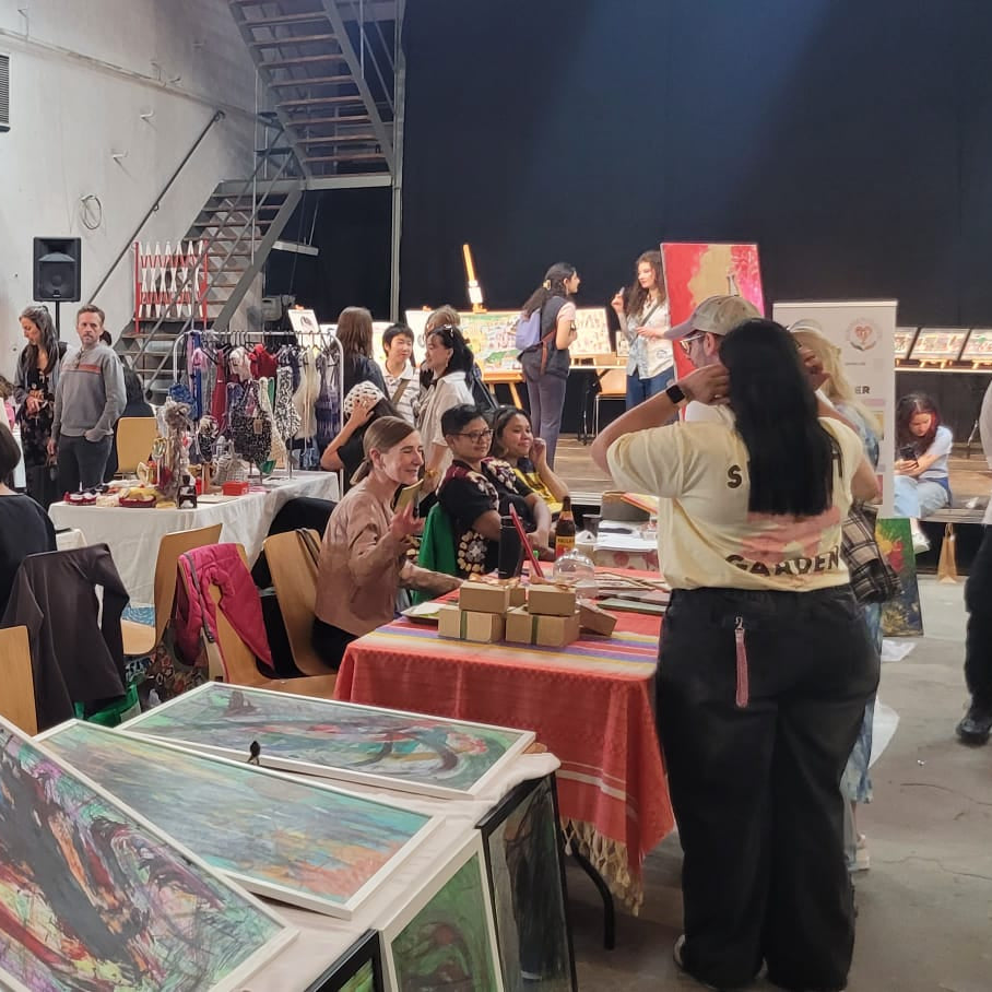 A crowded indoor arts and culture festival, fair or market with many people, tables, and displayed artwork. In the foreground, framed paintings or prints are shown on a table, with people walking and interacting throughout the space.