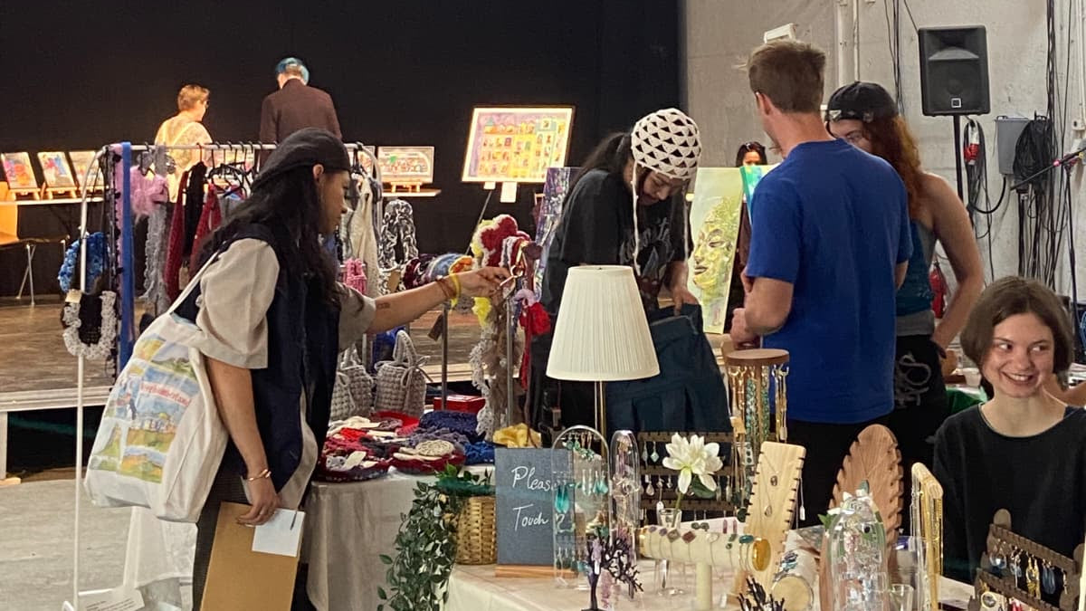 A bustling indoor market or craft fair scene in a large warehouse space with high ceilings and stage lighting. Several people are standing around and browsing tables displaying handmade items, including a white table lamp, glassware, and other decor.